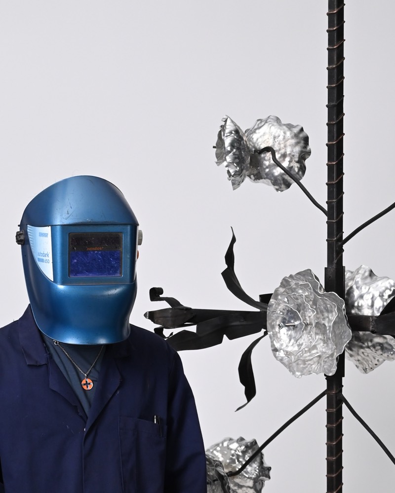 Daoud Qureshi in a blue welding mask and boiler suit beside a mild-steel sculpture of aluminium flowers on a rebar stem.