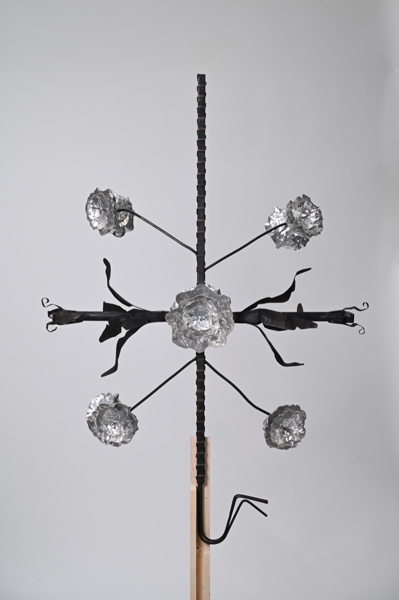 Full view of a mild-steel sculpture: a rebar stem carrying radially arranged aluminium blooms and forged black leaves.