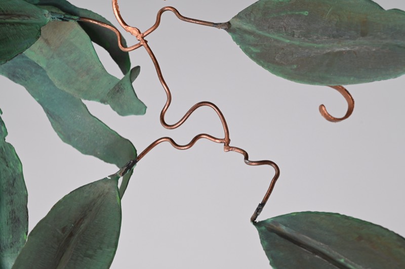 Detail of scrolled copper wire tendrils threading between patinated leaves.