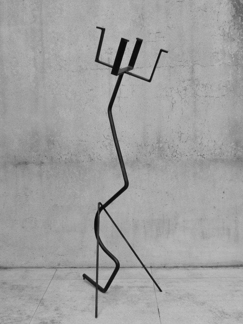 Full view of a gestural forged mild-steel figure standing against a raw concrete wall.