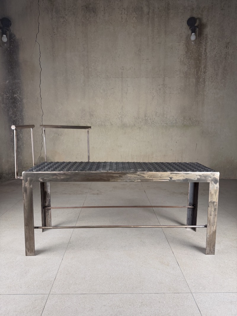 Front view of a raw mild-steel bench with a chequer-plate seat and thin rail back, in a bare concrete room.